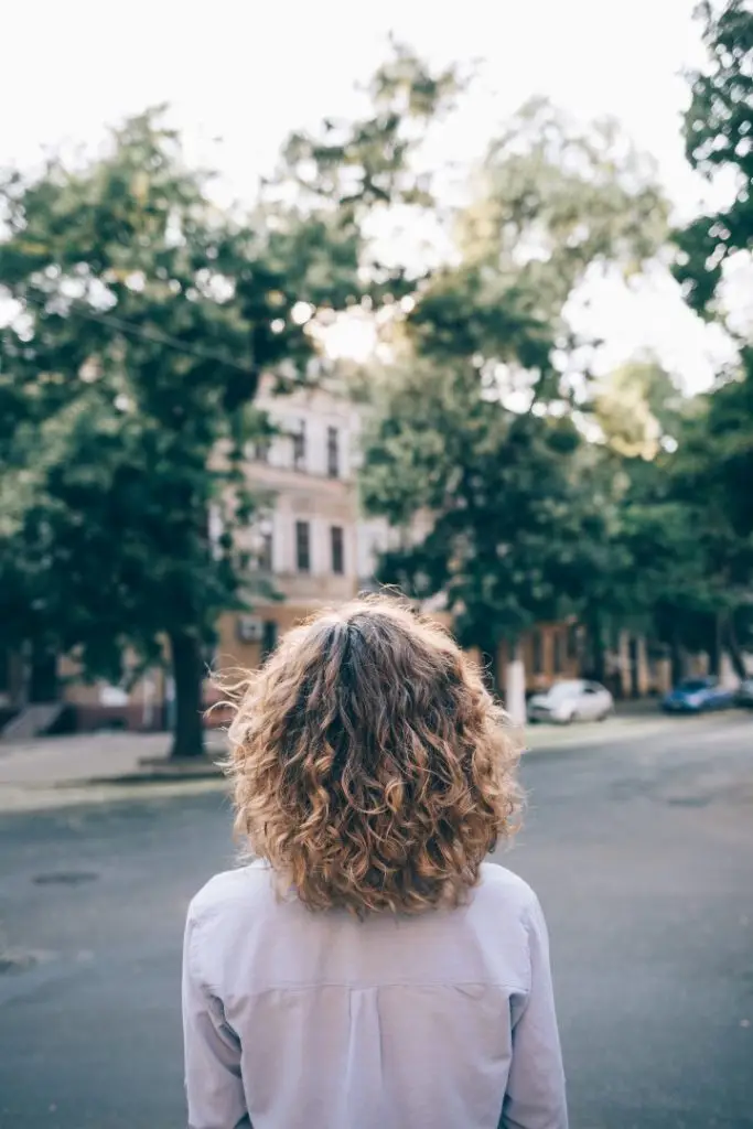 Young woman with thick curly hair looking at the street while walking around the city. Rear view of unrecognizable female wearing blue shirt standing outside on summer day.