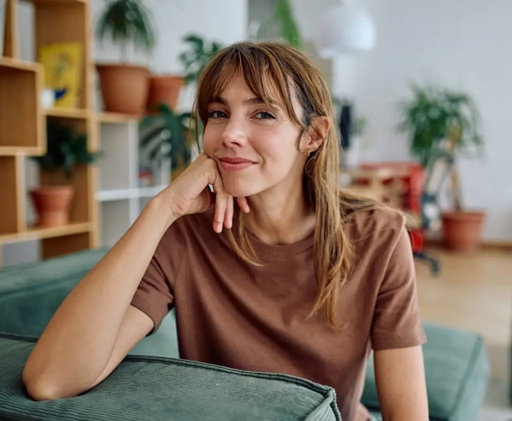 Portrait of a serene woman resting her chin on her hand while sitting on a comfortable sofa in her modern, plant-filled living room