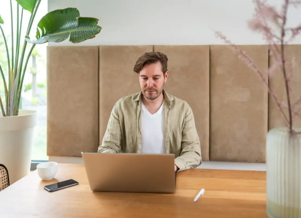A man diligently operates a laptop at a dining table, his casual attire blending seamlessly into the residential setting, highlighting the adaptability of modern workspaces.